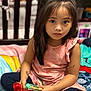 bedroom, black_pants, blanket, casual_clothing, child, colorful, cross_legged, curious_expression, doll, facial_expression, girl, indoors, long_hair, pink_top, playtime, sitting, soft_lighting, tattoo, toy, white_socks