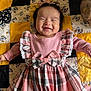 apparel, baby, bow, cheerful, child, cute, dress, fabric, face, happy, headband, indoors, infant, lying_down, mickey_mouse, pattern, pink, plaid, quilt, smiling