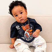 Jesus is registered to the contest to win money with this photo: toddler, child, curly_hair, couch, beige, navy_blue_tshirt, bear_graphic, ripped_pants, white_socks, sitting, indoors, cute, portrait, casual_clothing, young_child, gold_chain, expression, curious, comfortable, home