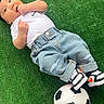 baby, infant, child, grass, soccer_ball, footwear, sneakers, jeans, white_shirt, hat, outdoor, sports, cute, lying_down, toy, green_background, casual, relaxed, portrait, young