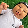 baby, infant, grass, greenery, white_shirt, nike_logo, beanie, black_hat, cute, smiling, happy, outdoor, portrait, child, small_hands, casual_clothing, lying_down, young_child, expressive_face, playful
