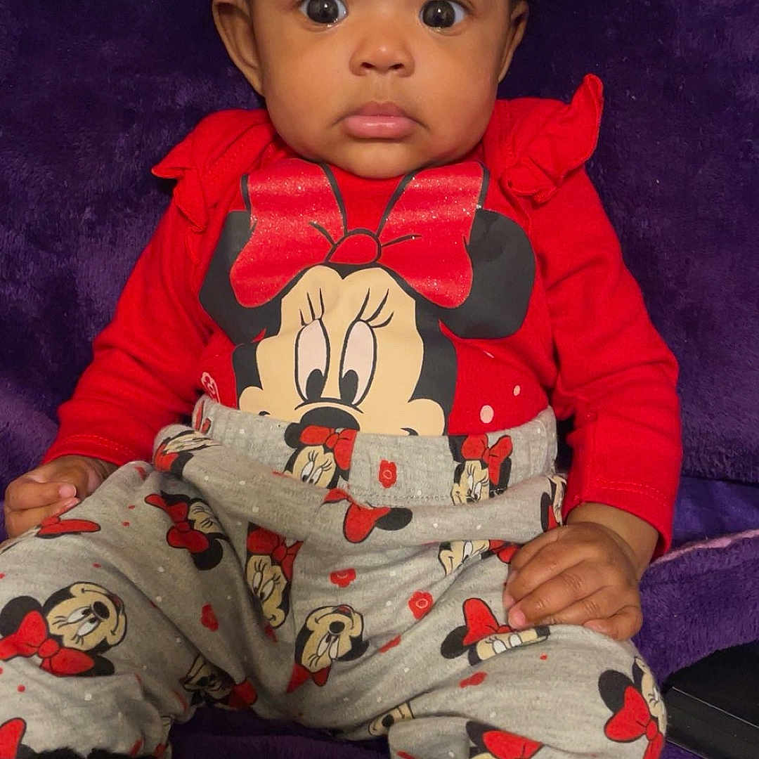 Ahmyra is registered to the contest to win money with this photo: apparel, baby, black_socks, child, cozy, cute, expression, face, hands, indoors, minnie_mouse, patterned_pants, person, plush_background, portrait, purple_background, red_clothing, sitting, wide_eyes, young_child