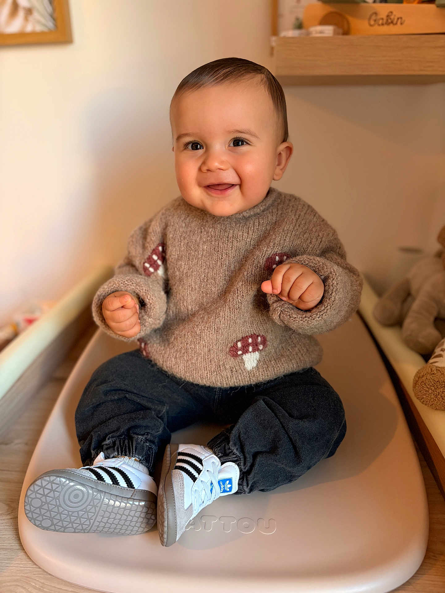 Gabin participe au concours pour gagner de l'argent avec cette photo : baby, child, smiling, sweater, mushroom_pattern, shoes, adidas, sneakers, pants, sitting, indoor, changing_pad, wooden_floor, toy, stuffed_animal, face, hands, happy, portrait, cozy