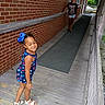 child, children, smiling, outdoor, ramp, walkway, brick_wall, wooden_floor, railing, blue_bow, dress, shorts, sandals, portrait, siblings, standing, happy, residential_building, greenery, daylight