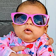 Aurora is registered to the contest to win money with this photo: baby, infant, child, sunglasses, pink_sunglasses, portrait, close_up, face, outdoor, colorful_clothing, strawberry_pattern, floral_pattern, onesie, supported, adult_hand, fashion, cute, photography, brick_background, door