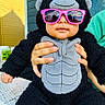 baby, child, infant, gorilla_costume, costume, sunglasses, pink_sunglasses, portrait, sitting, outdoor, humor, cute, fashion, playful, furry, black_costume, gray_clothing, adult_hands, patio, daylight