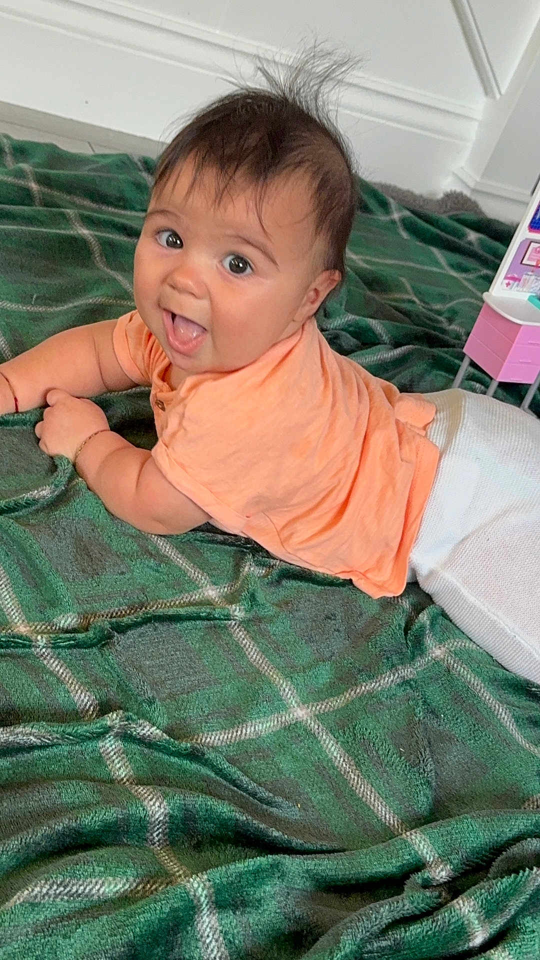 Thiago is registered to the contest to win money with this photo: baby, child, curious, cute, expression, face, floor, green_blanket, hair, happy, indoors, infant, lying_down, peach_shirt, person, plaid, playful, skin, smiling, white_pants