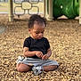 toddler, child, playground, wood_chips, sitting, black_shirt, gray_shorts, sneakers, curly_hair, outdoor, daytime, focused, play_area, equipment, nature, casual_clothing, young_child, person, kids, leisure