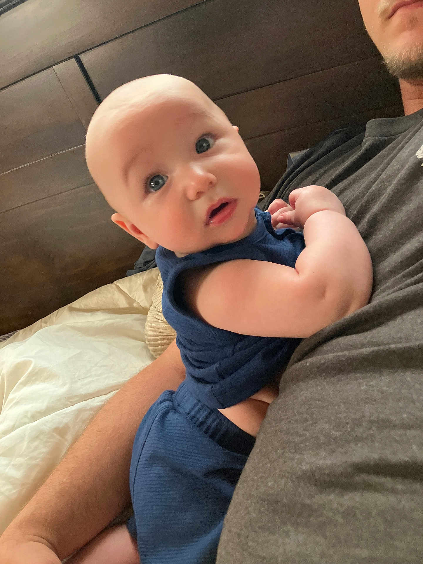 Levi joined the competition — help win amazing prizes! baby, infant, blue_eyes, navy_outfit, adult_arm, bed, pillow, wooden_headboard, skin, closeup, curious, indoor, comfort, cuddling, expression, person, child, portrait, soft_light, casual_clothing