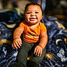 baby, infant, child, smiling, sitting, portrait, indoors, orange_shirt, blanket, star_pattern, dark_pants, bracelet, happy, grinning, short_hair, bed, soft_lighting, bokeh, close_up, photography
