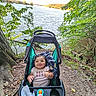 baby, child, stroller, lake, outdoor, nature, forest, water, trail, hiking, smiling, sitting, infant, toy, blanket, trees, leaves, shoreline, family_outing, portrait