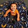 baby, child, infant, smiling, happy, sitting, portrait, face, curly_hair, orange_shirt, dark_pants, blanket, stars, moons, space_theme, indoors, closeup, warm_lighting, patterned_fabric, ai_generated