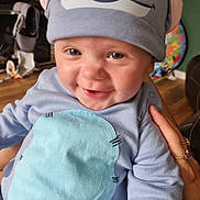 Logan is registered to the contest to win money with this photo: baby, child, smiling, blue_hat, clothing, indoor, person, hand, toy, furniture, floor, skin, face, eyes, mouth, happy, cute, infant, apparel, holding