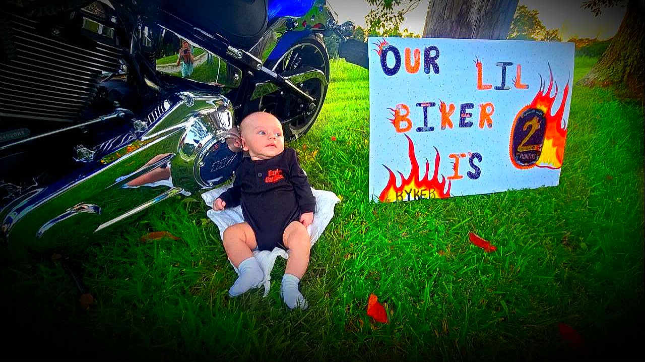 Ryker is registered to the contest to win money with this photo: baby, infant, motorcycle, black_clothing, grass, outdoor, sign, handmade_sign, text, harley_davidson, socks, chrome, reflection, tree, person, two_months, cute, child, motorbike, greenery