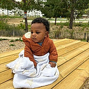 Khalil a rejoint le concours — aidez-le/la à gagner de superbes lots ! baby, child, outdoor, picnic_table, blanket, jacket, wood, nature, tree, greenery, park, fence, curly_hair, infant, seated, daylight, casual_clothing, expression, young, fall
