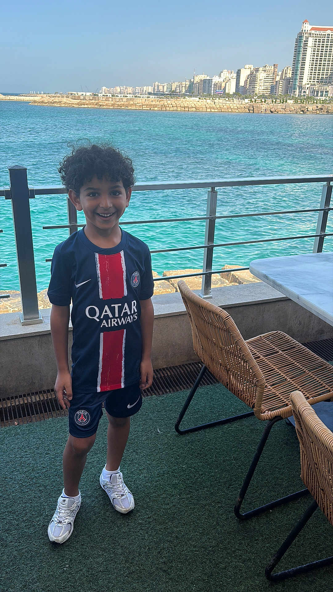 Zayn a rejoint le concours — aidez-le/la à gagner de superbes lots ! child, boy, smiling, sportswear, shorts, sneakers, curly_hair, waterfront, ocean, railing, cityscape, buildings, outdoor, daylight, patio, chair, table, grass_carpet, happy, person