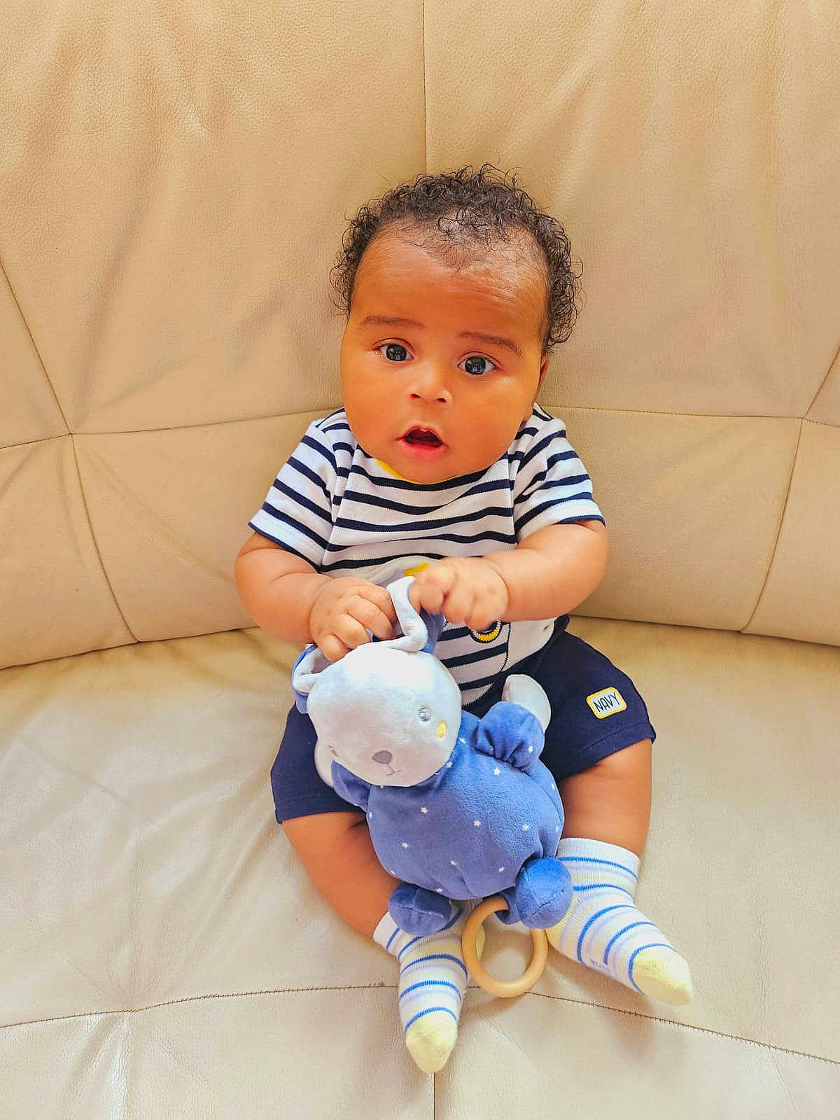 Sebastien-Michel a rejoint le concours — aidez-le/la à gagner de superbes lots ! baby, child, toy, blue_bunny, striped_shirt, navy_shorts, socks, couch, beige, curly_hair, sitting, indoor, cute, infant, plush_toy, person, young_child, expression, holding, soft_toy