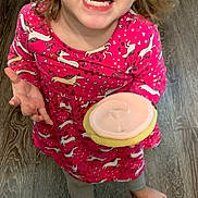 Riot is registered to the contest to win money with this photo: child, toddler, cookie, pink_dress, unicorn_pattern, barefoot, wooden_floor, happy, smiling, indoors, long_sleeve, clothing, person, cute, food, frosting, young_child, looking_up, standing, casual