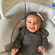 Irie is registered to the contest to win money with this photo: baby, smiling, infant, baby_seat, gray_clothing, headband, happy, child, portrait, indoor, cushion, tiled_floor, person, cute, young_child, baby_girl, closeup, comfortable, hand, face