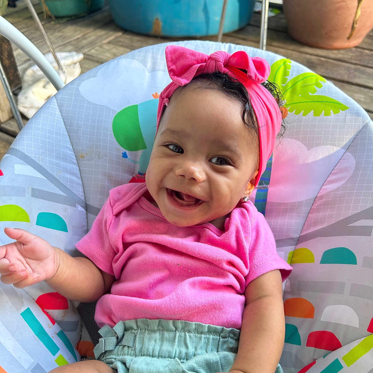 Kamari is registered to the contest to win money with this photo: baby, child, smiling, pink_headband, pink_shirt, green_skirt, baby_seat, colorful, outdoor, wooden_floor, potted_plants, happy, infant, portrait, cute, clothing, seat, person, headwear, playful