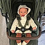 baby, stroller, child, smile, hood, seatbelt, socks, happy, indoor, carpet, brown_pants, jacket, child_seat, infant, person, baby_clothing, footwear, baby_smiling, baby_in_stroller, baby_sitting