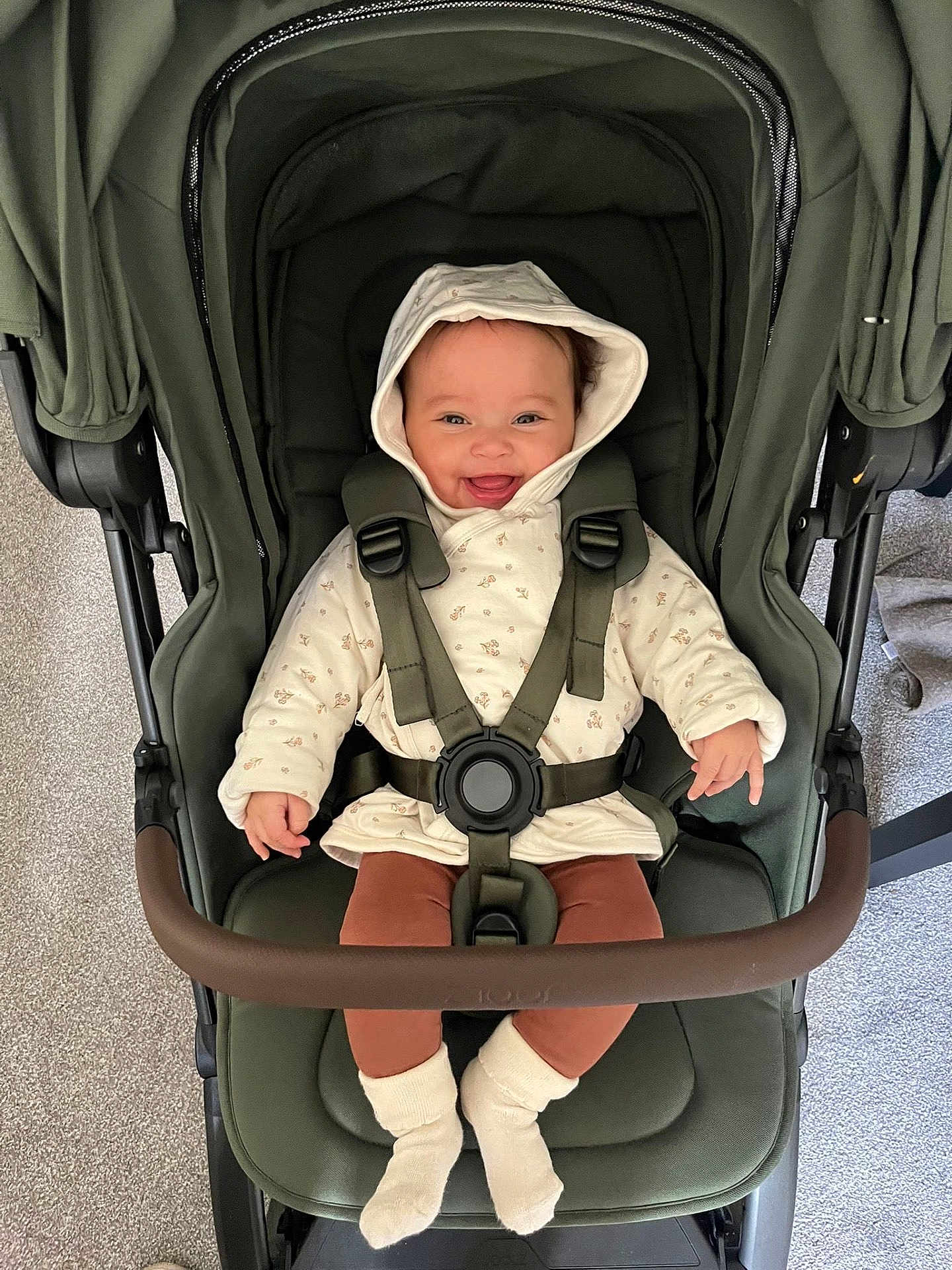 Ariella is registered to the contest to win money with this photo: baby, stroller, child, smile, hood, seatbelt, socks, happy, indoor, carpet, brown_pants, jacket, child_seat, infant, person, baby_clothing, footwear, baby_smiling, baby_in_stroller, baby_sitting