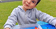 Kevin is registered to the contest to win money with this photo: blue_table, casual_clothing, child, curious, daylight, face, fruit, grass, hoodie, nature, outdoor, park, plastic_container, playful, portrait, raspberries, smiling, strawberries, trees, young_child