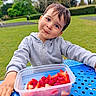 Kevin is registered to the contest to win money with this photo: blue_table, casual_clothing, child, curious, daylight, face, fruit, grass, hoodie, nature, outdoor, park, plastic_container, playful, portrait, raspberries, smiling, strawberries, trees, young_child