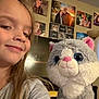 child, smile, selfie, stuffed_animal, plush_cat, toy, living_room, family_photos, wall_art, furniture, lamp, blond_hair, casual_clothing, happy, indoors, portrait, cute, home, decor, childhood