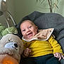 baby, smiling, yellow_sweater, bib, plush_toy, glowing_toy, gray_blanket, cozy, indoor, child, happy, cute, infant, soft_toy, portrait, seated, warm_light, toy_bear, candid, comfort