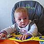 baby, blue, book, child, clothing, curious, expression, face, hands, high_chair, indoor, infant, orange, person, portrait, seatbelt, table, toy, yellow, young_child