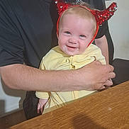 Lou participe au concours pour gagner de l'argent avec cette photo : baby, child, person, adult, hand, arm, face, smile, headband, red_horns, yellow_clothing, table, wood, indoor, window, black_shirt, seated, holding, happy, cute