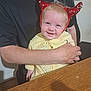 baby, child, person, adult, hand, arm, face, smile, headband, red_horns, yellow_clothing, table, wood, indoor, window, black_shirt, seated, holding, happy, cute