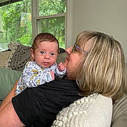 Theodore joined the competition — help win amazing prizes! baby, woman, indoor, window, couch, blanket, pillow, glasses, dinosaur_print, curly_hair, child, holding, portrait, smile, person, home, cozy, natural_light, family, affection