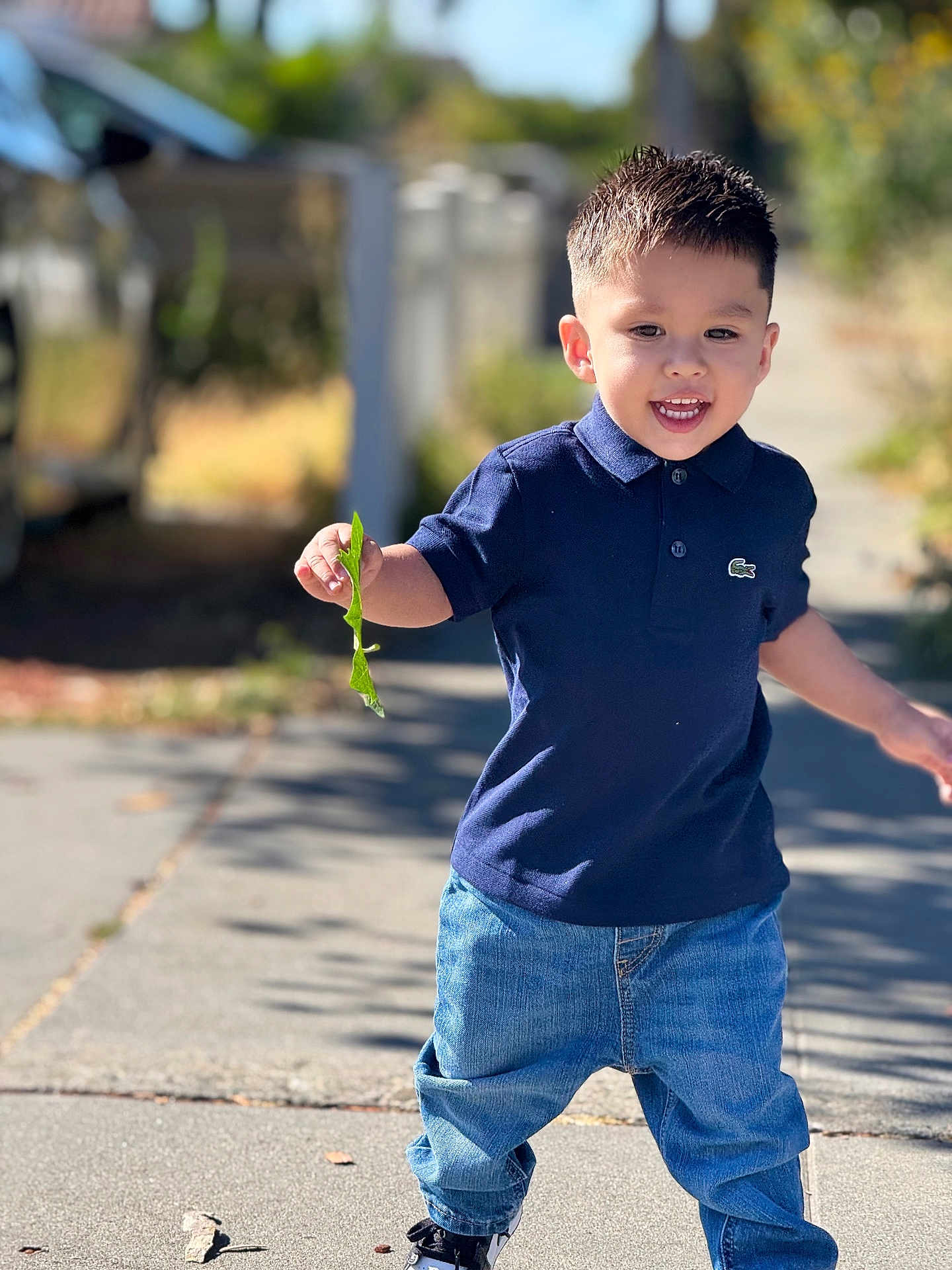 Joel Hernandez is registered to the contest to win money with this photo: child, toddler, boy, smiling, outdoor, sidewalk, leaf, blue_polo, jeans, casual_clothing, daylight, sunlight, happy, walking, short_hair, nature, fence, shoes, young_child, playful