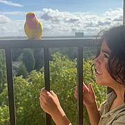 Ayden participe au concours pour gagner de l'argent avec cette photo : child, bird, balcony, bars, yellow_bird, red_bird, curly_hair, outdoor, daylight, greenery, sky, clouds, nature, happy, smiling, person, holding, curious, sunlight, portrait