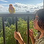 child, bird, balcony, bars, yellow_bird, red_bird, curly_hair, outdoor, daylight, greenery, sky, clouds, nature, happy, smiling, person, holding, curious, sunlight, portrait