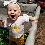 Miley joined the competition — help win amazing prizes! baby, child, playpen, indoor, smiling, happy, white_bow, white_shirt, yellow_shorts, carpet, furniture, fan, blanket, green_blanket, snowflake_pattern, person, standing, infant, cute, room