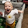baby, child, playpen, indoor, smiling, happy, white_bow, white_shirt, yellow_shorts, carpet, furniture, fan, blanket, green_blanket, snowflake_pattern, person, standing, infant, cute, room