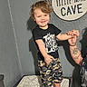 Monty is registered to the contest to win money with this photo: child, toddler, adult, smiling, holding_hands, indoor, wall, sign, text, cushion, camouflage_shorts, black_tshirt, tattoo, happy, person, portrait, curly_hair, playful, family, interaction