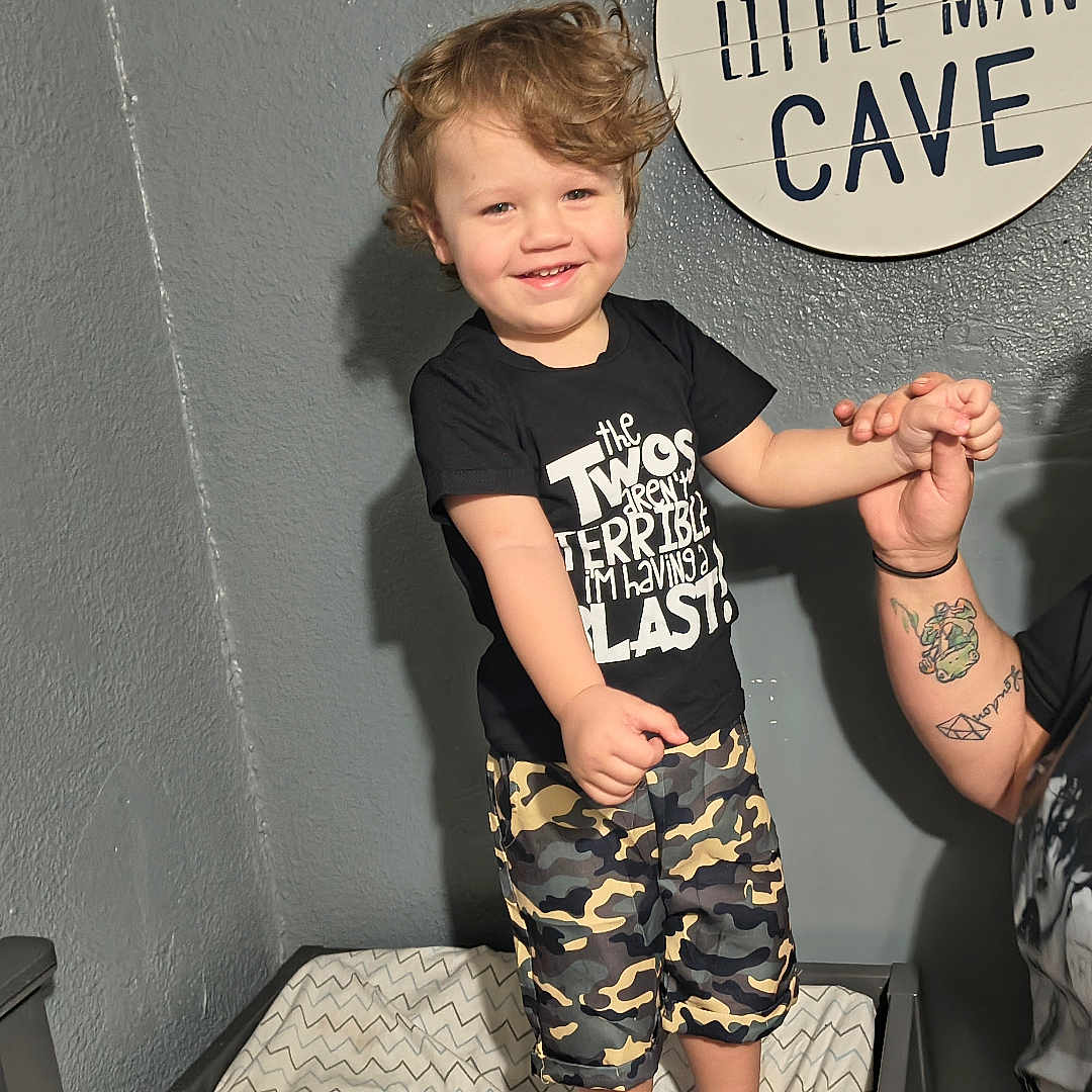 Monty is registered to the contest to win money with this photo: child, toddler, adult, smiling, holding_hands, indoor, wall, sign, text, cushion, camouflage_shorts, black_tshirt, tattoo, happy, person, portrait, curly_hair, playful, family, interaction