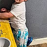 toddler, child, curly_hair, standing, play_kitchen, yellow_table, blue_crocs, shorts, white_tshirt, indoor, wood_floor, gray_wall, smiling, looking_back, young_child, casual_clothing, home, cute, playtime, portrait