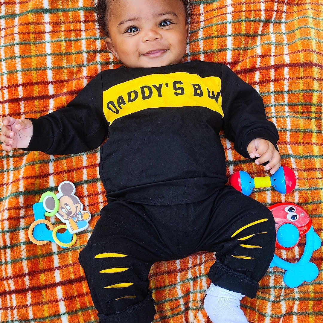 Romari is registered to the contest to win money with this photo: baby, black_clothing, cheerful, child, colorful, curly_hair, cute, happy, indoors, infant, lying_down, mickey_mouse_toy, plaid_blanket, playful, portrait, rattle, smiling, toy, white_socks, yellow_text