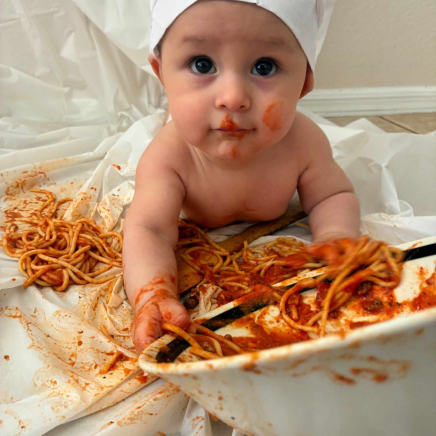 Elias is registered to the contest to win money with this photo: baby, child, spaghetti, chef_hat, messy, food, sauce, playing, curious_eyes, face_paint, cooking, cuisine, indoor, white_background, plate, hand, expression, cute, infant, toddler