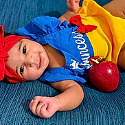 Nicolle is registered to the contest to win money with this photo: baby, child, smiling, headband, red_bow, blue_outfit, yellow_skirt, apple, lying_down, portrait, cute, infant, indoor, person, happy, closeup, skin, hand, face, blue_background