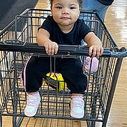 Raniah is registered to the contest to win money with this photo: baby, child, toddler, shopping_cart, grocery_store, indoor, portrait, face, curly_hair, black_clothing, sneakers, pink_shoes, sitting, gripping, handlebar, wooden_floor, retail, shopping, serious_expression, store_aisle