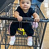 baby, child, toddler, shopping_cart, grocery_store, indoor, portrait, face, curly_hair, black_clothing, sneakers, pink_shoes, sitting, gripping, handlebar, wooden_floor, retail, shopping, serious_expression, store_aisle