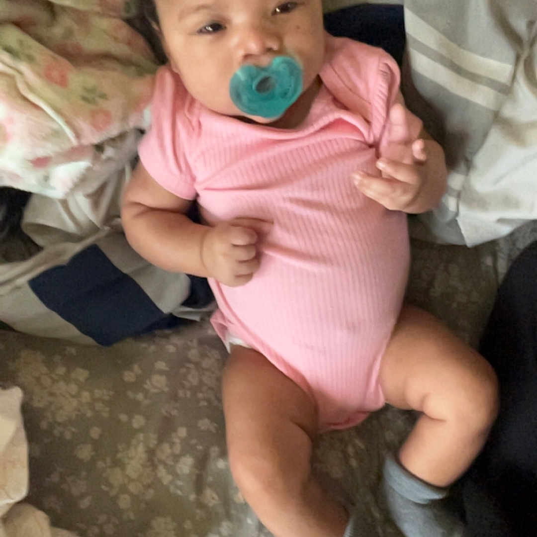 Raniah joined the competition — help win amazing prizes! baby, pacifier, pink_onesie, gray_socks, bed, blanket, pillow, infant, child, lying_down, indoors, person, cute, sleepy, soft_lighting, casual, cozy, resting, portrait, young_child