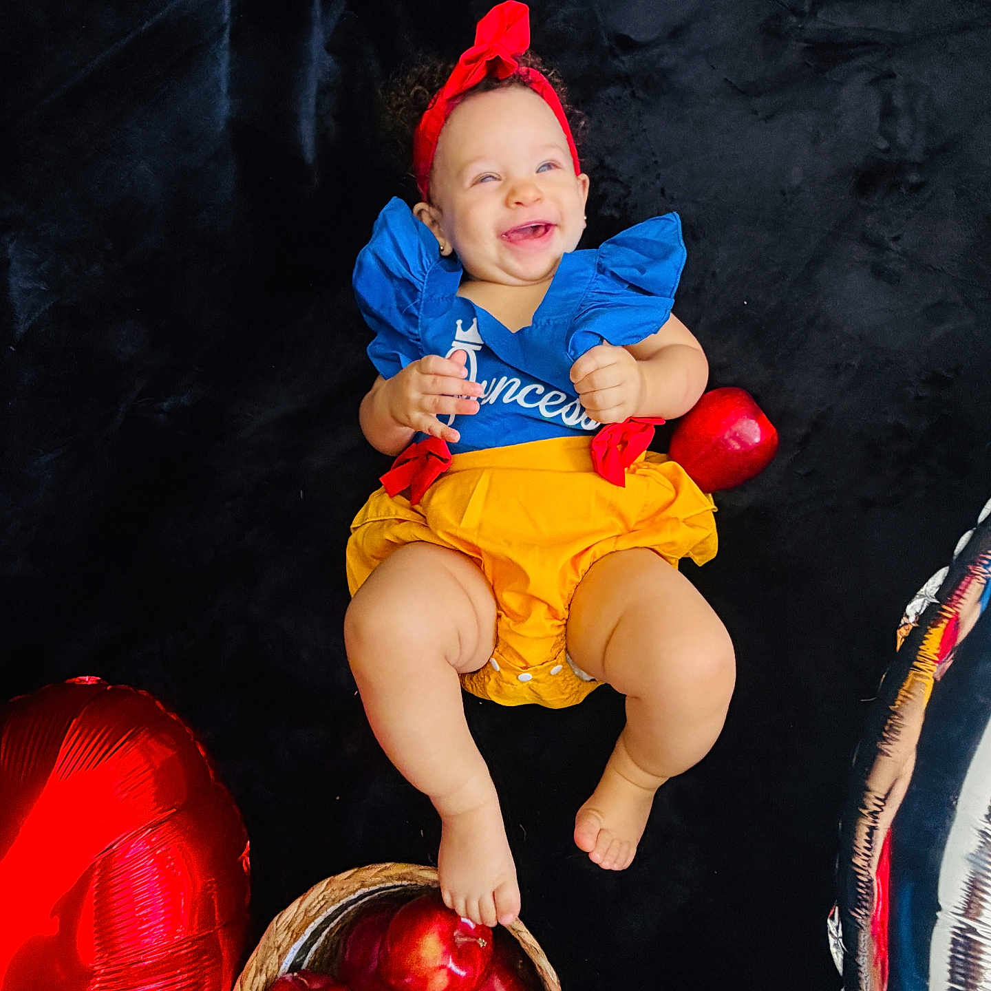 Aliyah Olivia is registered to the contest to win money with this photo: baby, child, infant, smiling, happy, portrait, costume, princess, headband, blue_dress, yellow_skirt, red_balloons, silver_balloon, apples, fruit_basket, indoor, black_background, sitting, celebration, photography