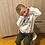 child, boy, smiling, happy, pose, hoodie, white_clothing, cargo_pants, green_pants, blue_shoes, sneakers, sitting, stool, wooden_floor, cabinet, indoors, playful, casual, portrait, person